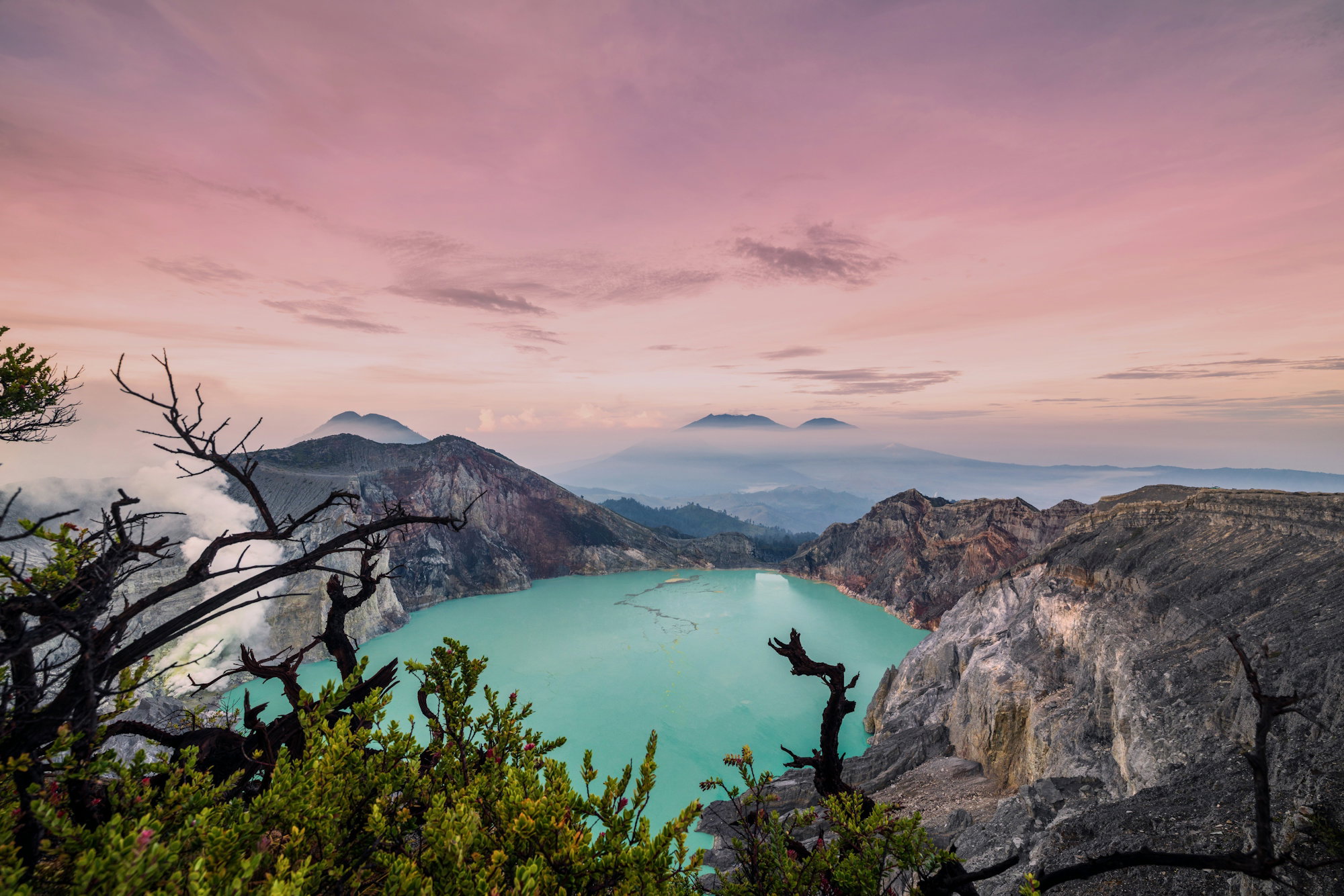 A turquoise volcanic crater lake surrounded by rocky cliffs under a soft pink sky